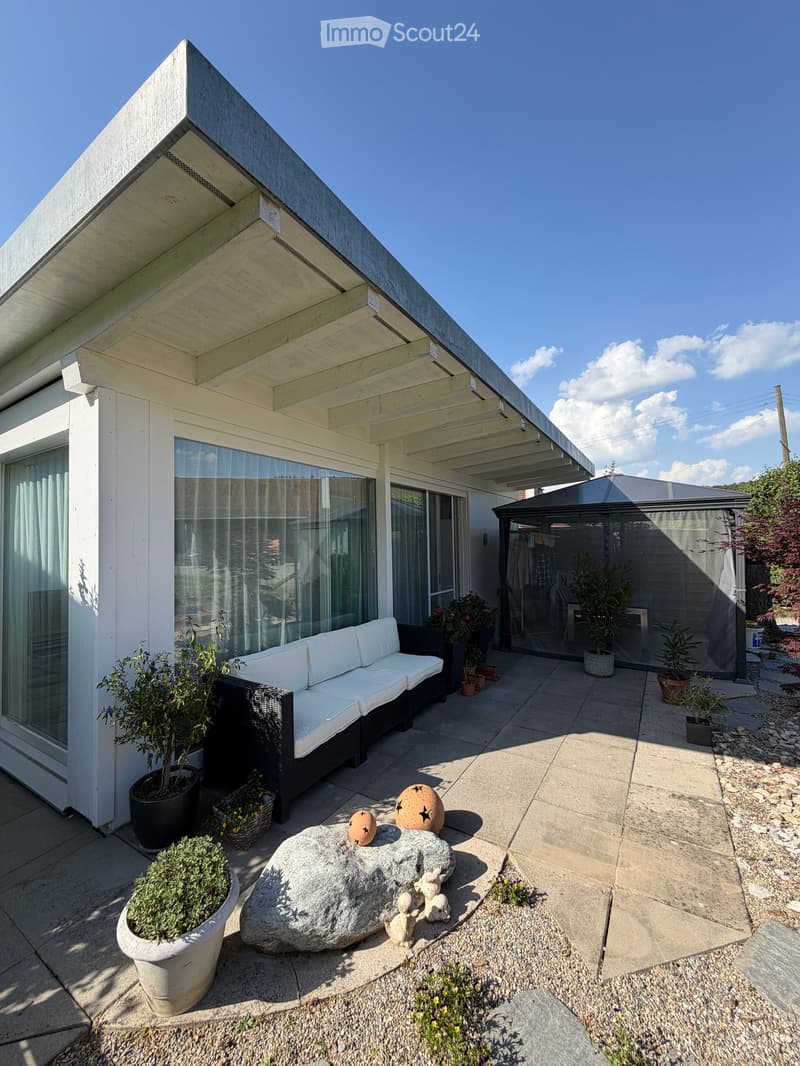 Modern outdoor patio with a white sofa, decorative rock, potted plants, and a covered seating area with glass walls.