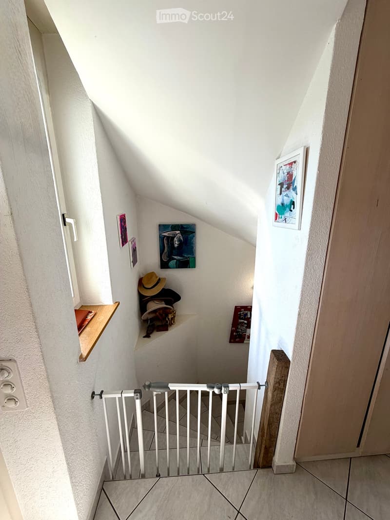 A narrow, slanted hallway with white walls, tiled floor, and a white safety gate at the bottom. Shelves on the walls hold hats, small items, and framed artwork.