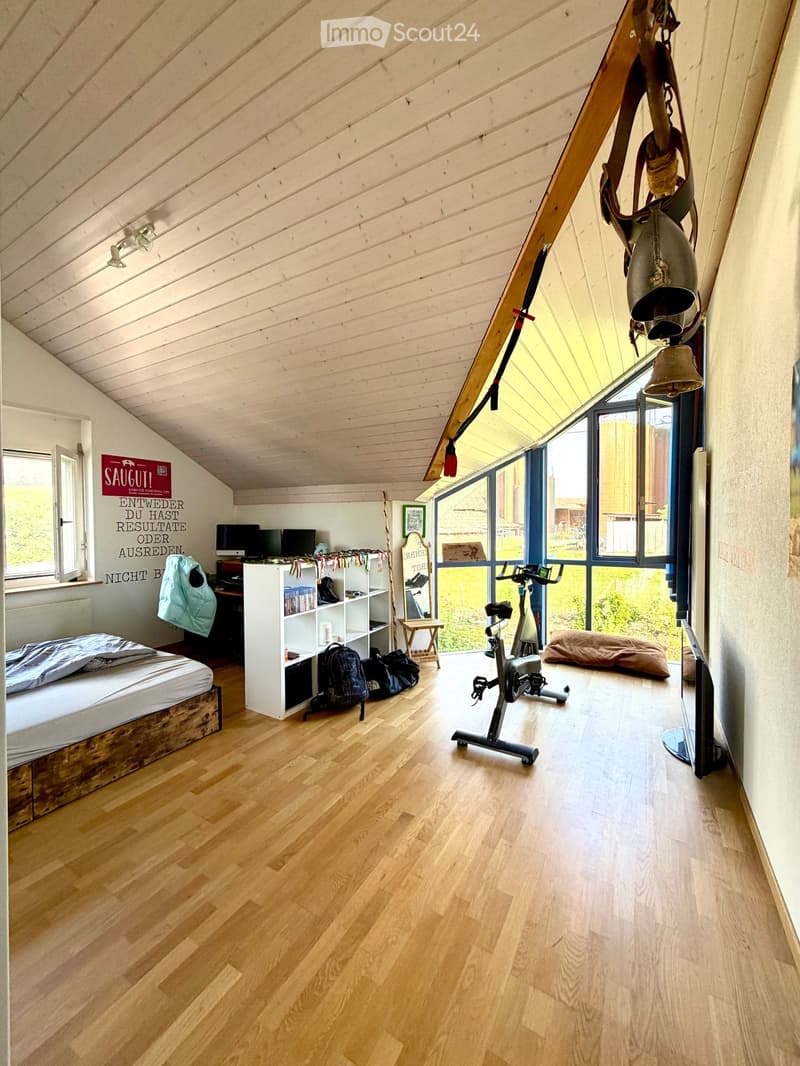 Loft-style bedroom with wooden floors, slanted wooden ceiling, large windows, bed, shelf, exercise bike, and screen.