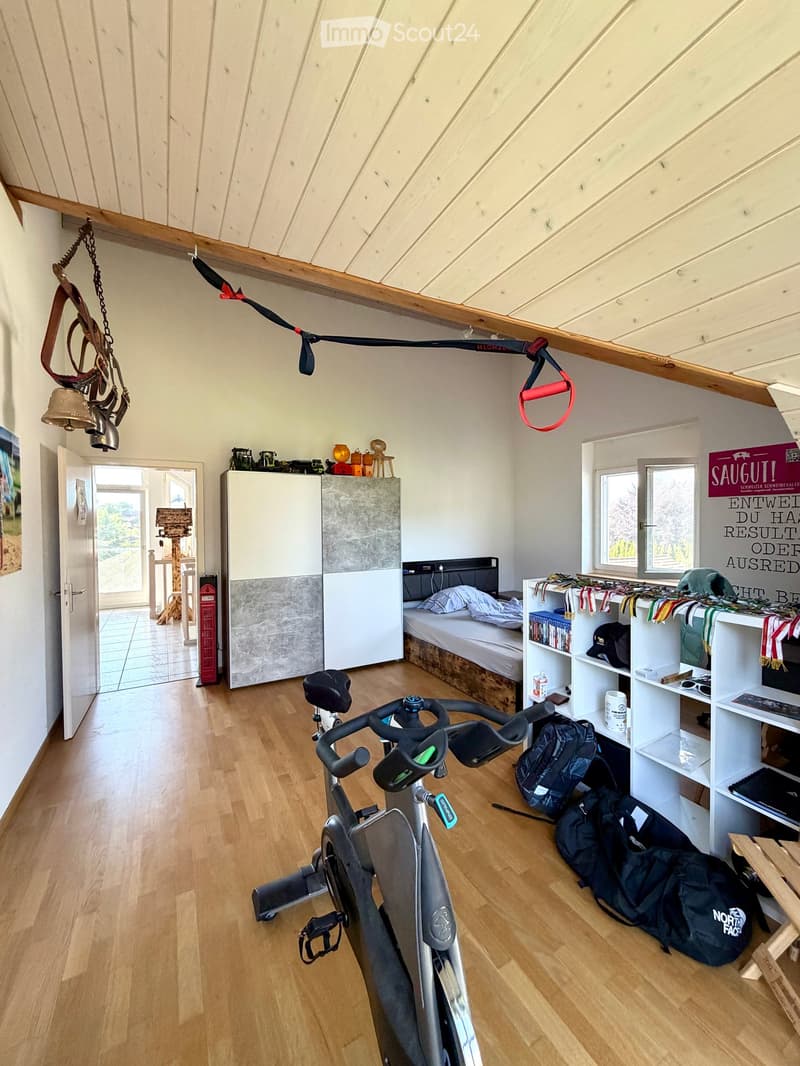 A multi-purpose room with wooden flooring, an exercise bike, a bed, shelves with books and bags, and a cabinet. The room has large windows and a door leading outside.