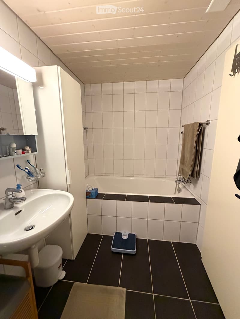 Modern bathroom with white tiles, a sink with a mirror cabinet, a bathtub, a towel on a rack, and a scale on the floor.