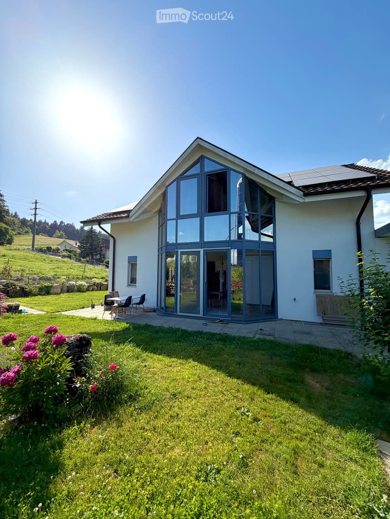 Modern two-story house with large glass windows and a spacious garden, featuring a patio area with outdoor furniture.