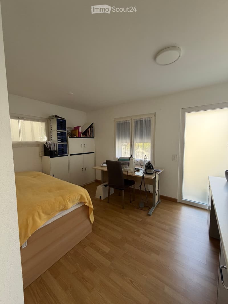 A well-lit bedroom with a wooden floor, a bed with a yellow blanket, a desk with a chair, a cabinet, and a storage unit. The room has large windows with curtains and a sliding glass door.