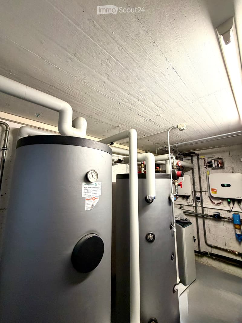 Utility room with two water heaters, pipes, valves, and a control panel mounted on the wall.