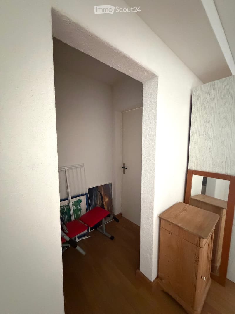 A small, narrow hallway with wooden flooring, white walls, a wooden cabinet, a mirror, and a red stool in the background.