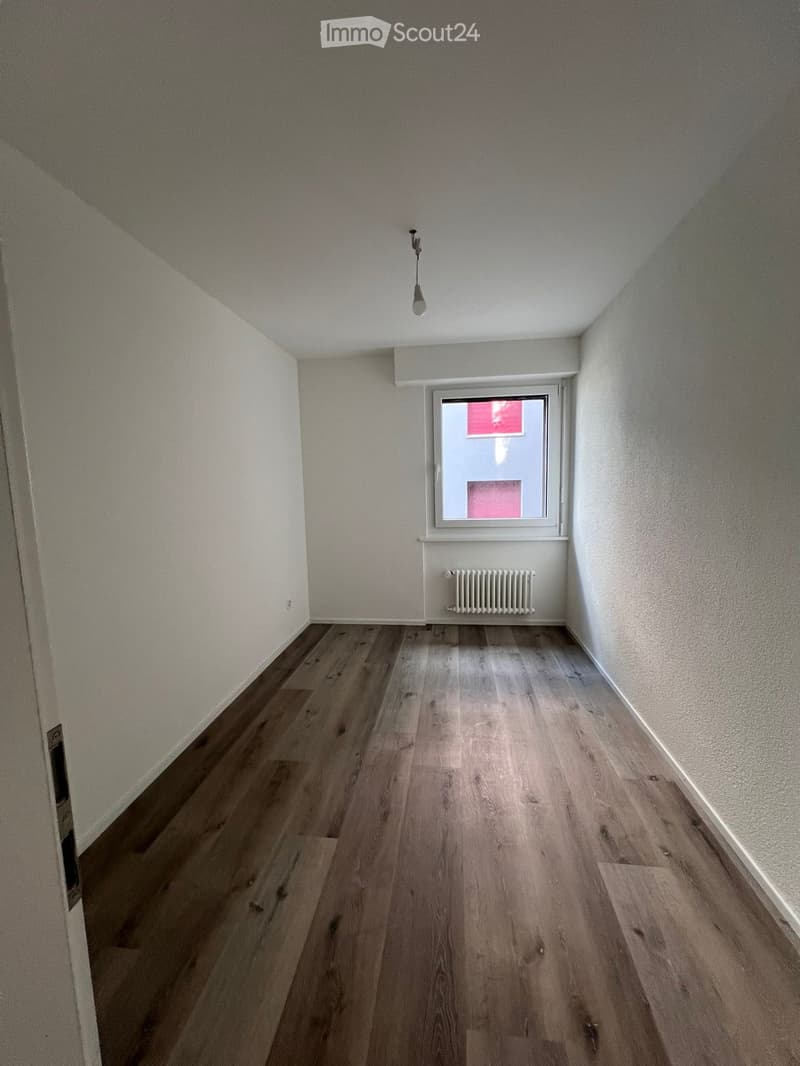 Empty room with wooden flooring, white walls, a single window with a radiator below, and a ceiling light fixture.