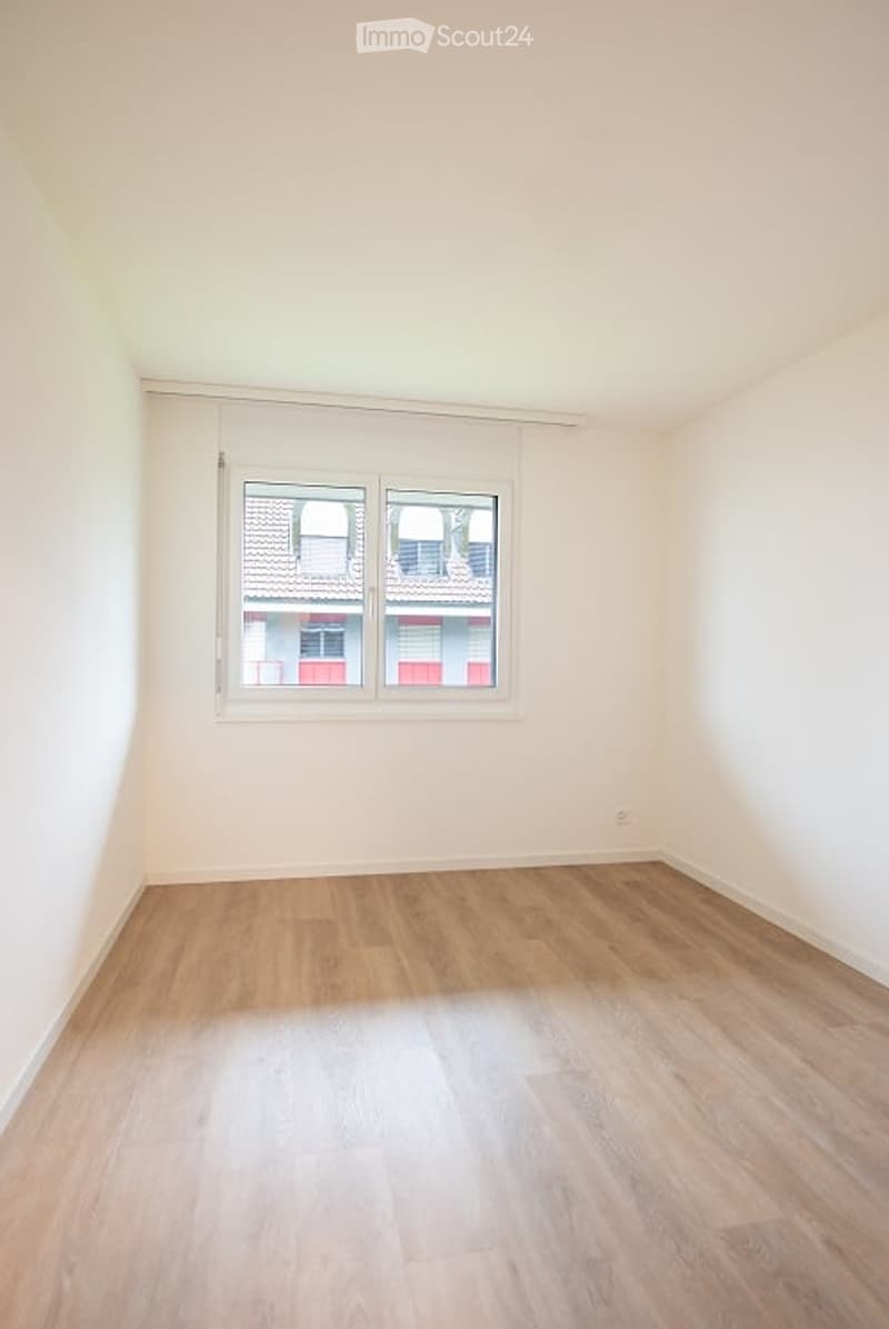 Empty room with light wooden flooring and white walls, featuring a large window with white frame and two panes, offering a view of neighboring buildings.