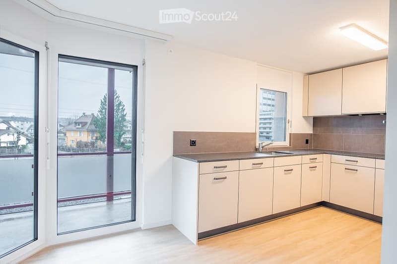 Empty kitchen with white cabinets, a sink, and a window overlooking a cityscape. The space features light wooden flooring and a sliding glass door leading to a balcony.
