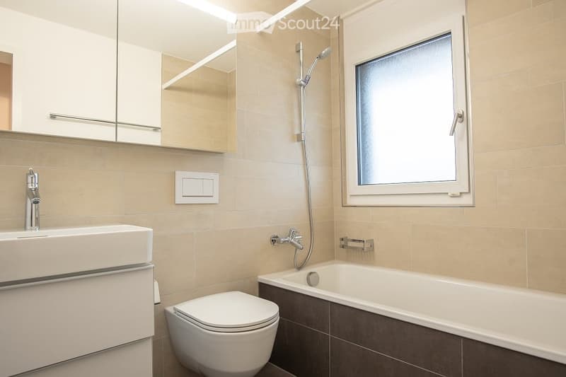 Modern bathroom with beige tiles, a white sink, toilet, bathtub, and a showerhead. Features a large mirror with built-in storage and a frosted window for privacy.