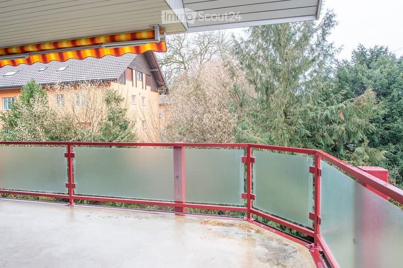 Balcony with red metal railing and glass panels, overlooking a residential area with trees and neighboring houses.