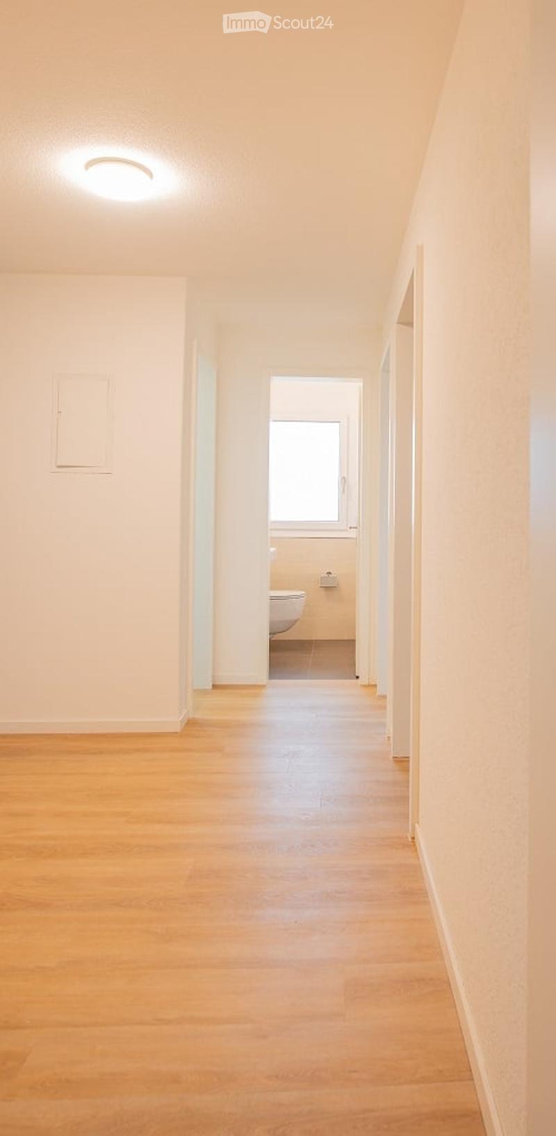 Empty hallway with light wooden flooring, white walls, and a recessed ceiling light. A partially visible bathroom is seen at the end of the hallway.