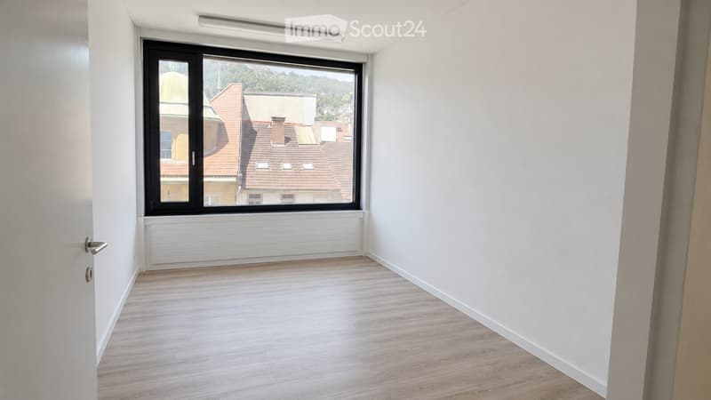 Empty room with light wooden flooring, white walls, and a large window offering a view of rooftops and greenery.