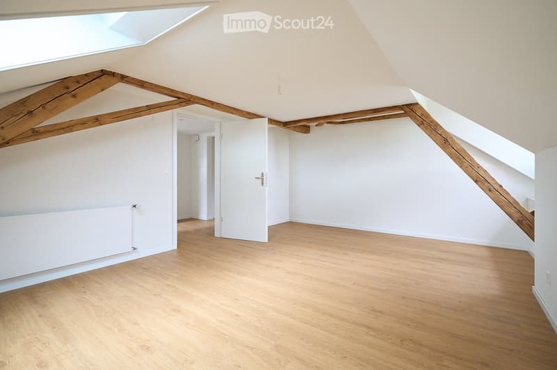 Empty attic room with wooden beams, skylight, and wooden flooring. No furniture or equipment present.