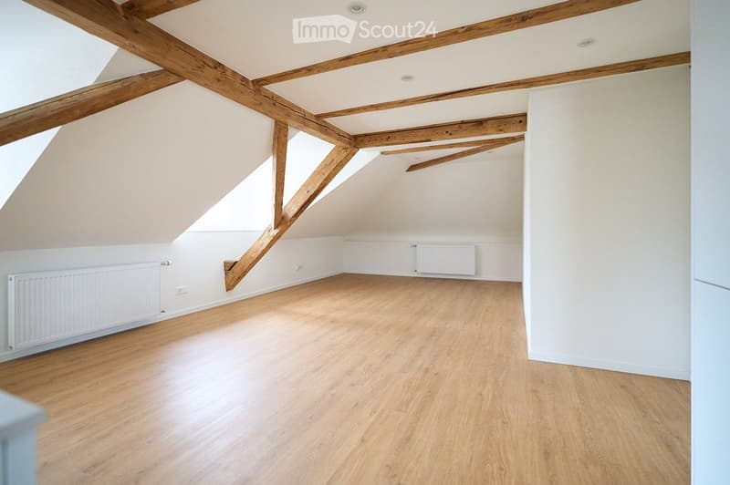 Empty attic room with wooden beams, wooden flooring, white walls, and radiators on two walls.