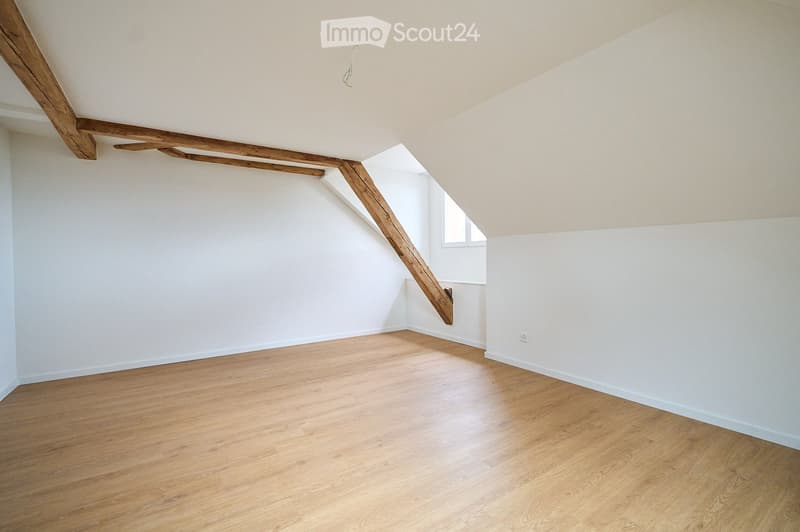 Empty room with wooden flooring, white walls, and exposed wooden beams on the ceiling. A small window is visible on one wall.