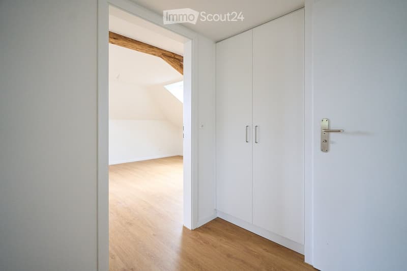 Empty room with light wooden flooring, white walls, and a built-in white closet with silver handles. The adjacent room has exposed wooden beams and a skylight.