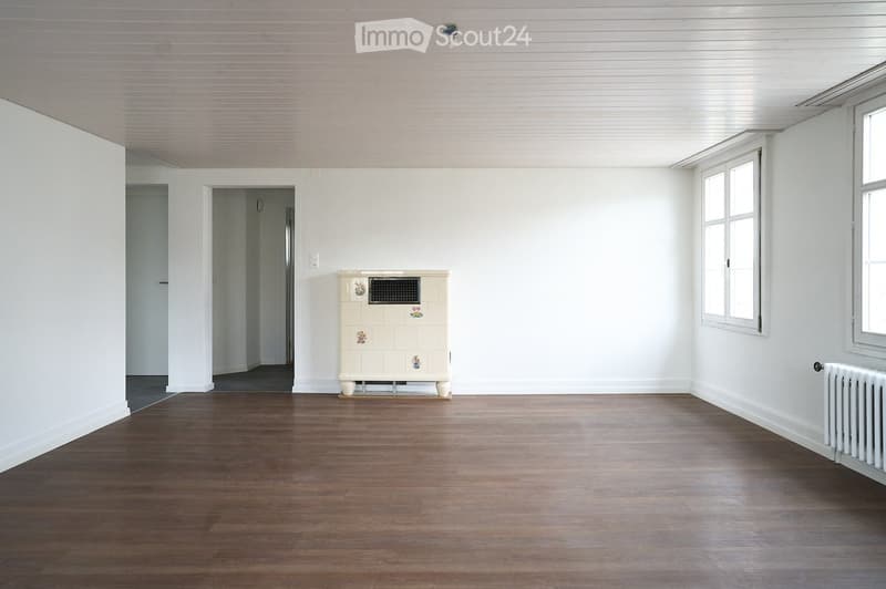 Empty room with wooden flooring, white walls, and large windows. Features a radiator and a decorative tiled cabinet.