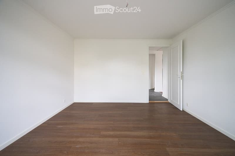 Empty room with white walls and wooden flooring, featuring an open doorway leading to another room.