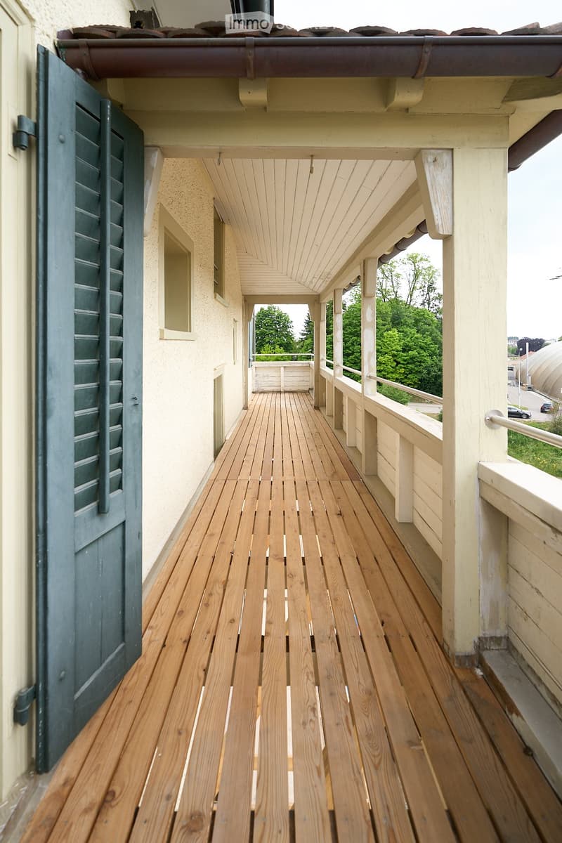 Wooden balcony with white railings, green shutters, and a view of greenery and a road.