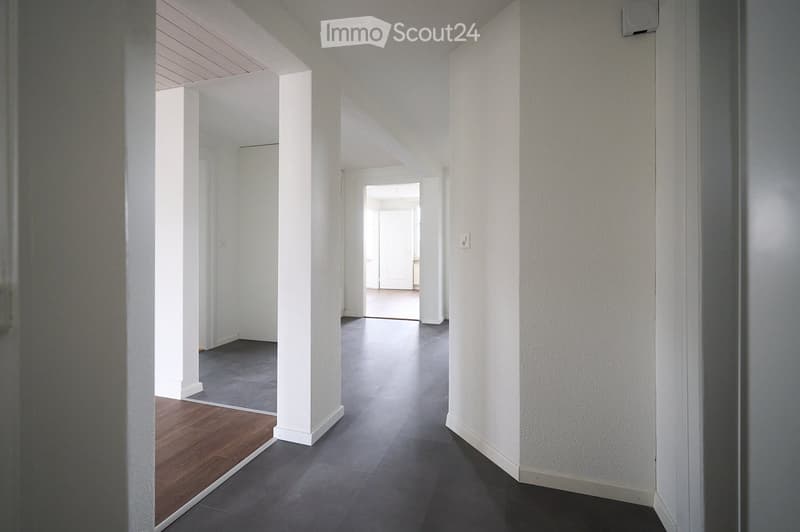 Empty hallway with white walls, dark grey tiled floor, and white columns. Natural light streams in from the left side, and a wooden floor is visible in the adjacent room.