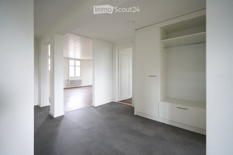 Empty room with white walls, grey tiled floor, built-in white closet with shelves and drawers, and a doorway leading to another room with a window and radiator.