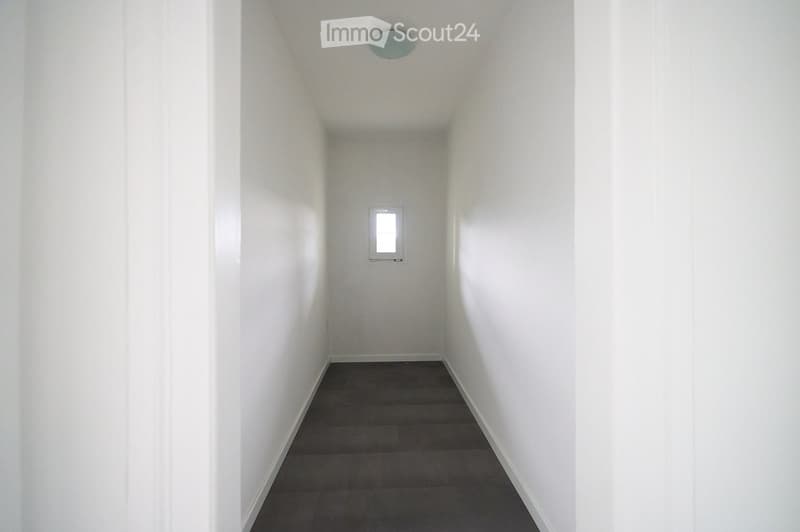 A narrow, empty hallway with white walls, a dark tiled floor, and a small window at the far end. The ceiling features a single light fixture.