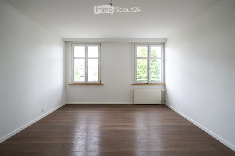 Empty room with wooden flooring, white walls, and two windows with wooden sills. A radiator is visible under one window.