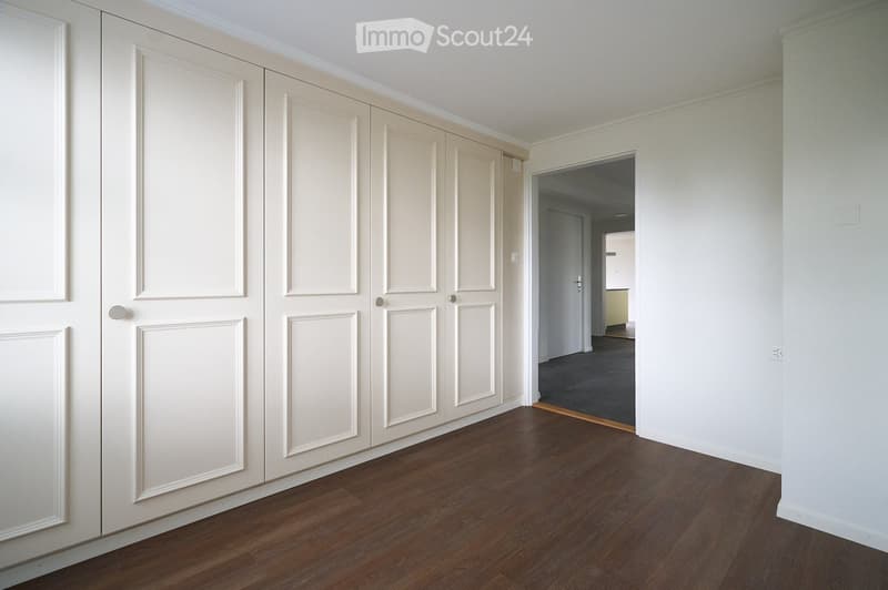 Empty hallway with wooden flooring, white walls, and a set of large white double doors on the left side. A doorway leads to another room with carpeted flooring visible in the background.