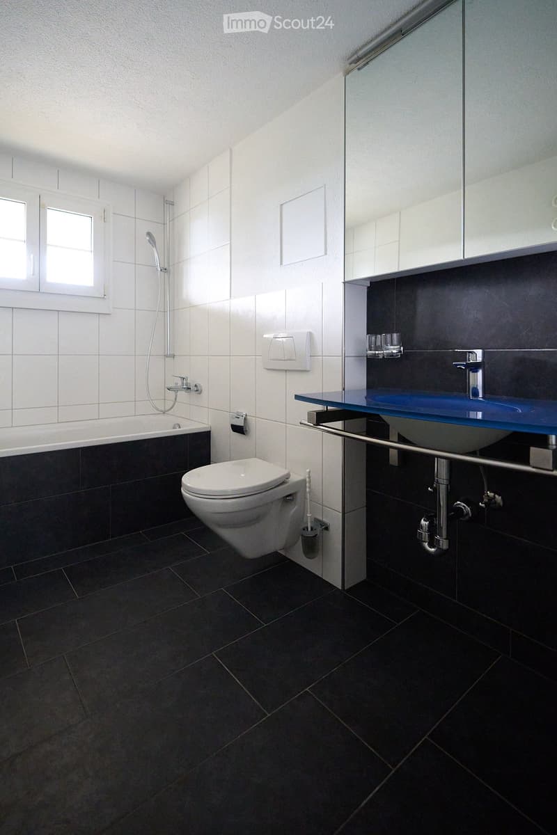 Modern bathroom with dark tiled floor, white tiled walls, bathtub, toilet, sink with blue countertop, and a large mirror above the sink.