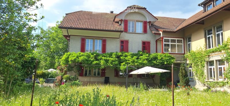 Two-story house with red shutters, beige walls, and a tiled roof. The house is surrounded by a lush garden with trees, flowers, and outdoor furniture.