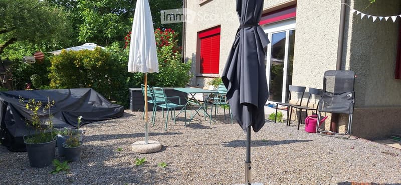 Garden area with gravel ground, patio furniture including a table, chairs, and umbrellas, along with potted plants and a covered object.