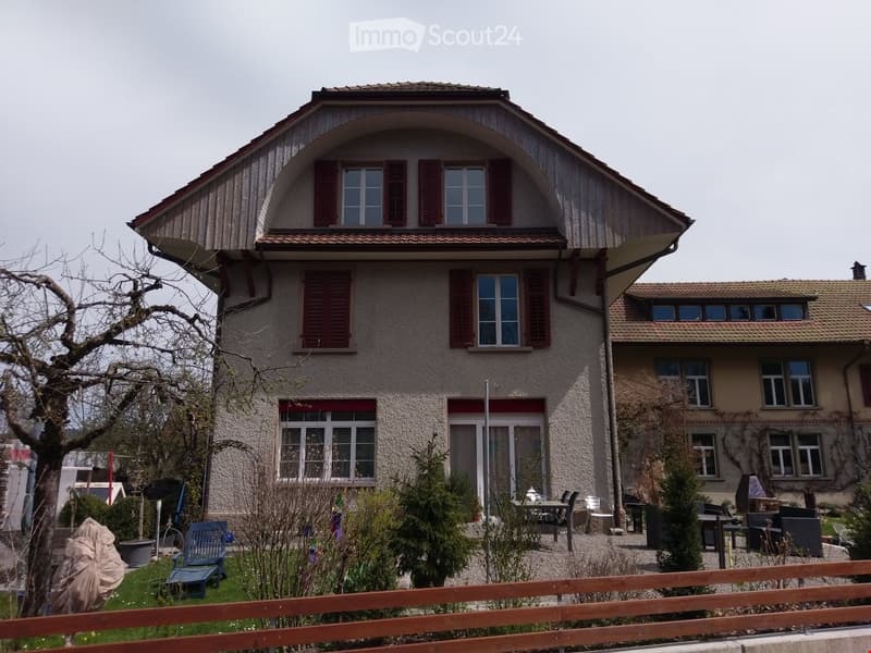 Two-story house with beige facade, red shutters, multiple windows, and a small garden area with outdoor furniture.