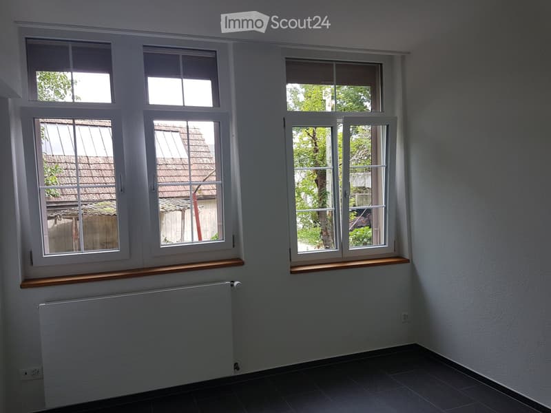 Empty room with white walls, dark tiled floor, multiple windows with wooden sills, radiator, and visible electrical outlets.