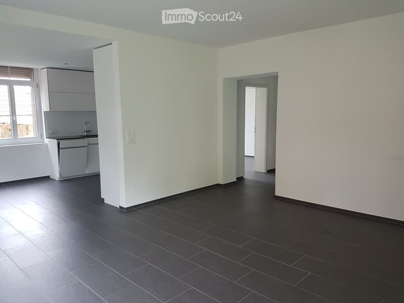 Empty room with white walls, dark tiled floor, and a small kitchen area with cabinets and sink visible in the background.