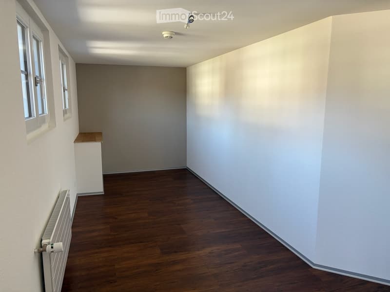 Empty room with white walls, wooden floor, and two windows. A small wooden shelf is against one wall, and a radiator is mounted on the left wall.