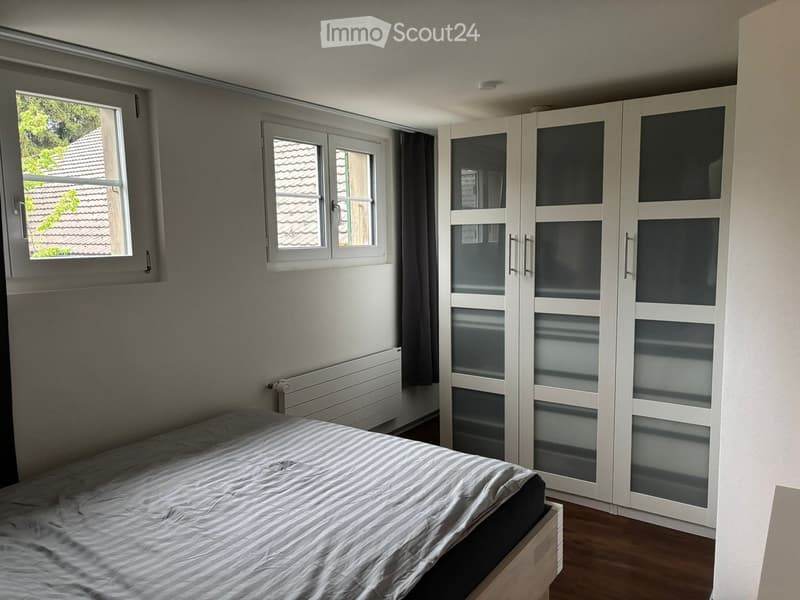 A bedroom with a bed, a wardrobe, and windows. The room has a wooden floor and white walls.