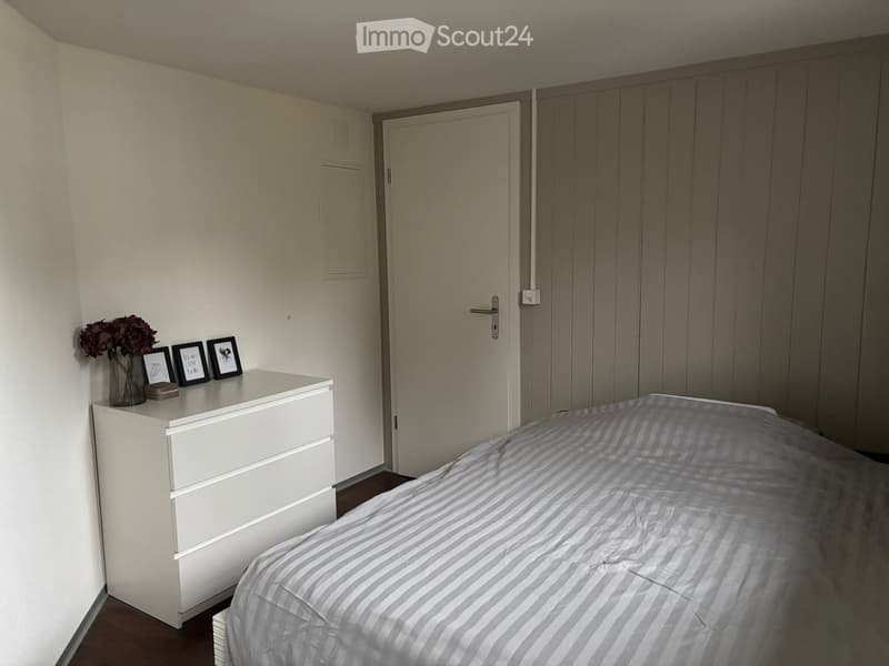 A bedroom with a bed, a dresser, and a vase with flowers on top. There are three picture frames on the dresser.