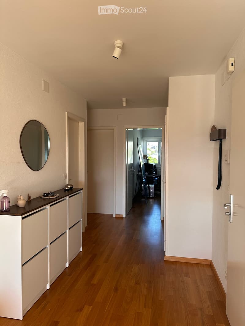 A clean, modern hallway with wooden flooring, white walls, and several doors leading to different rooms. A small console table with a mirror and some decorative items is visible on the left side.