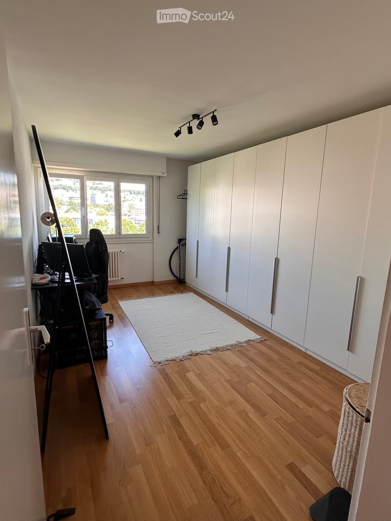Bright room with wooden flooring, large windows, white built-in cabinets, a small desk with a computer, and a white rug.