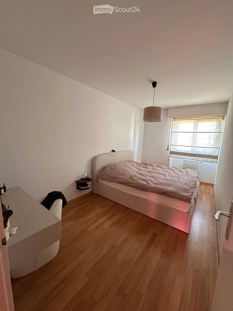 A small, modern bedroom with a wooden floor, a bed with a patterned duvet, a nightstand, a chair, and a hanging lamp.