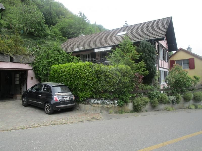 Two-story house with a tiled roof, surrounded by greenery and a small garden. A black car is parked in the driveway next to a garage entrance.