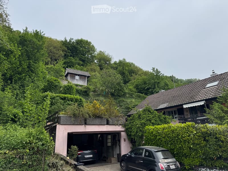 A two-story house with a garage, surrounded by lush greenery and trees. The garage has an open door with a car parked inside, and another car is parked outside. The house features a tiled roof and a small balcony on the upper level.