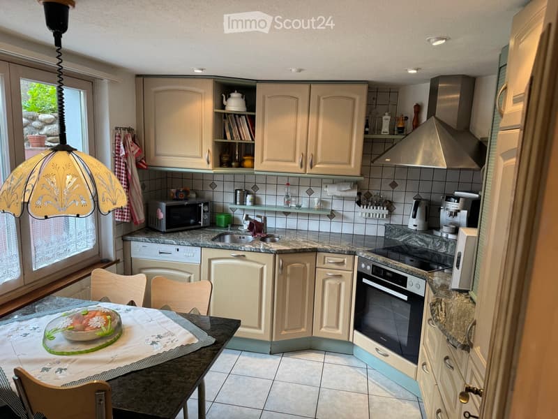 Well-equipped kitchen with cream-colored cabinets, granite countertops, and various appliances including an oven, microwave, dishwasher, and coffee maker. A dining area with a table and chairs is visible, along with a decorative lamp and a window with a v