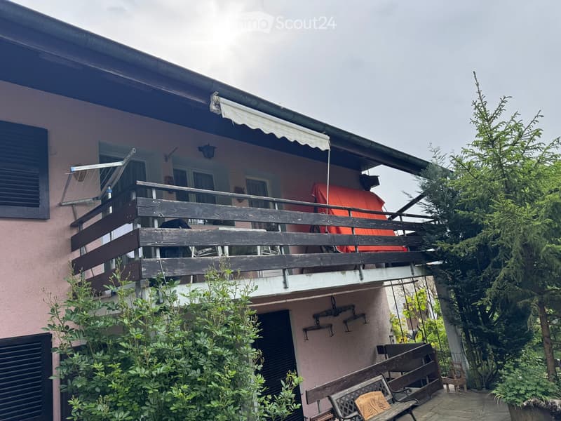 Two-story house with a balcony, wooden railing, and outdoor seating area. The balcony features a red cushioned chair and a small table. The house has a light pink exterior with multiple windows and a covered awning.