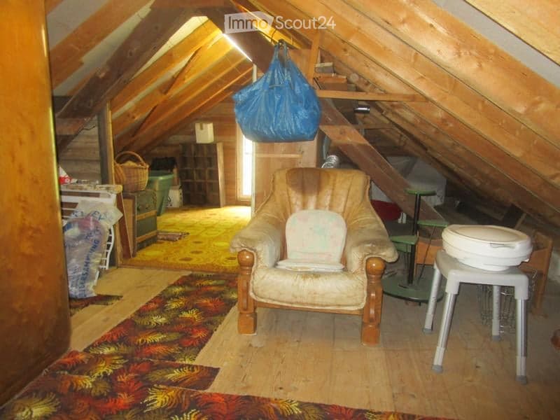 Attic room with wooden beams, slanted ceilings, and exposed wooden walls. Contains a leather armchair, a small table, a stool, a basket, and a plastic bag hanging from the ceiling.