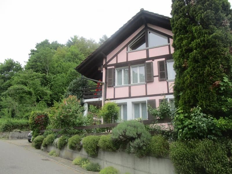 Two-story pink and brown house with a balcony, surrounded by greenery and a low stone wall.