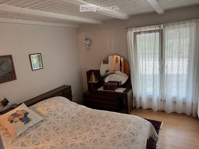 Cozy bedroom with a wooden bed, nightstand, dresser, mirror, and decorative items. Light-colored walls and wooden flooring.