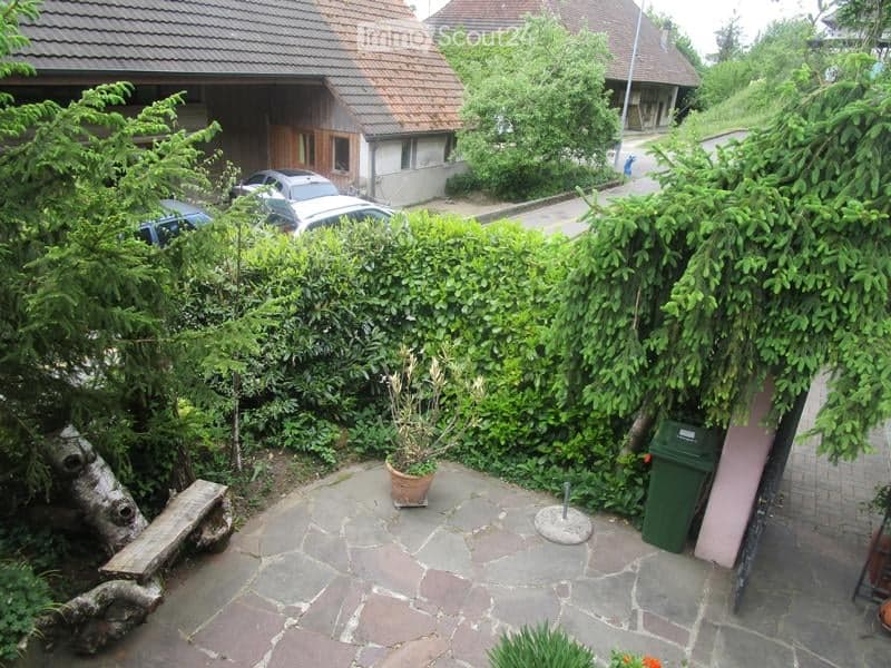 Garden area with stone patio, wooden bench, potted plants, and surrounding greenery. A house with a garage and parked cars is visible in the background.