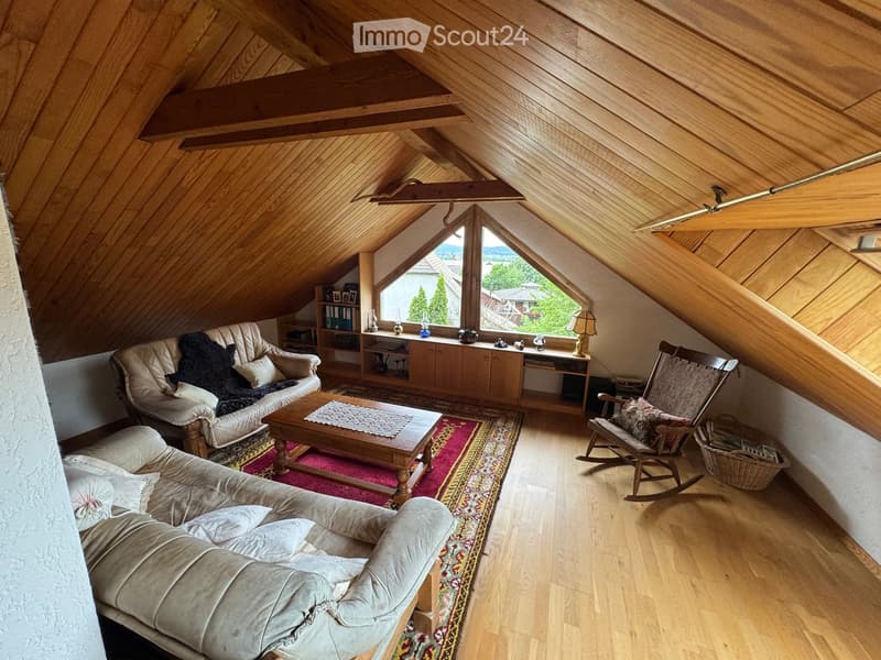 Cozy living room with wooden beams, slanted ceiling, large skylight window, sofa, coffee table, rocking chair, bookshelf, and table lamp.