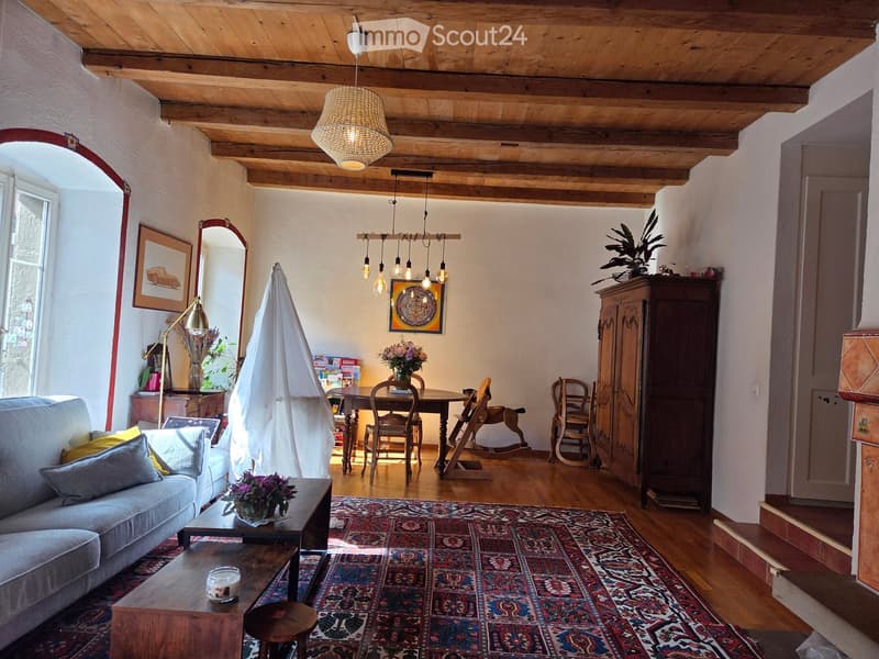A cozy living and dining area with exposed wooden beams, a large sofa, coffee table, dining table with chairs, a rocking horse, and decorative items.